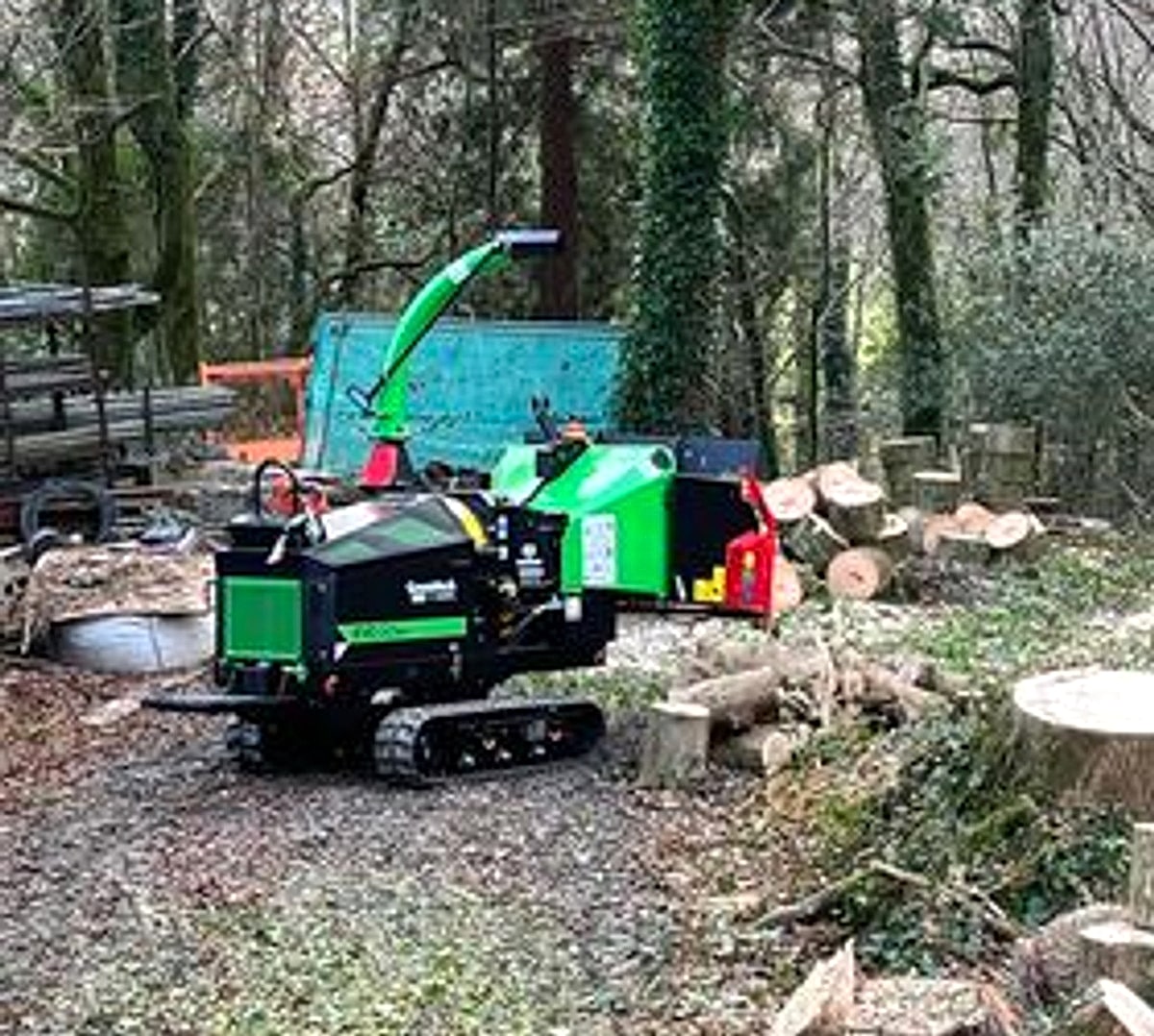 Tracked wood chipper and equipment in a wooded work area with logs nearby